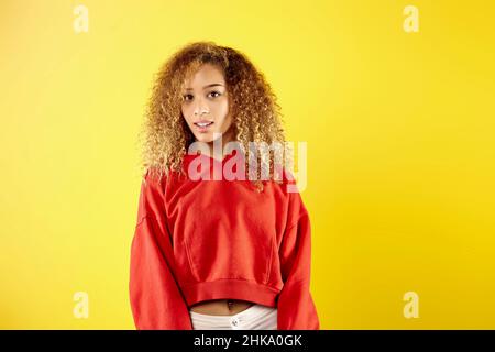 Confident young African American woman in stylish red hoodie and eyeglasses standing on yellow background in studio  Stock Photo