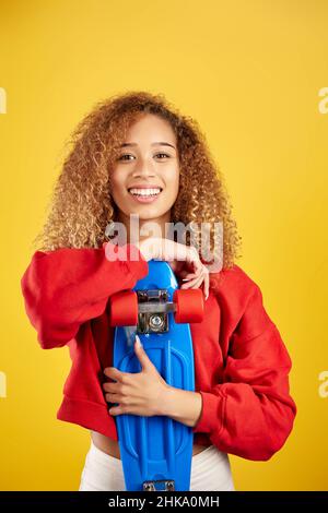 Delighted African American young female hipster in hoodie and with penny board looking at camera against yellow background Stock Photo