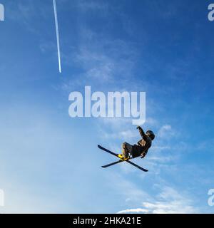 Ski jumper flying high in the air on a blue sky background with a trace of a jet aircraft. Stock Photo