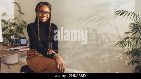 Young female freelancer smiling at the camera while working on a new project in her home office. Happy young photographer sitting on a chair with her Stock Photo