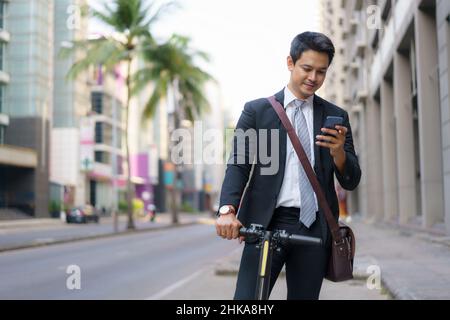 Asian businessman is riding an electric scooter and using his mobile phone to open a map in application to check the city road routes to work in the m Stock Photo