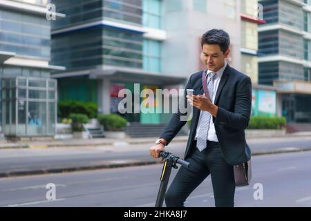 Asian businessman is riding an electric scooter and using his mobile phone to open a map in application to check the city road routes to work in the m Stock Photo