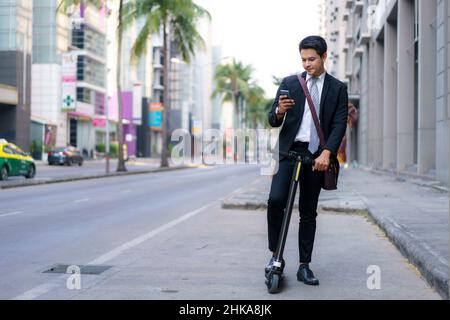Asian businessman is riding an electric scooter and using his mobile phone to open a map in application to check the city road routes to work in the m Stock Photo