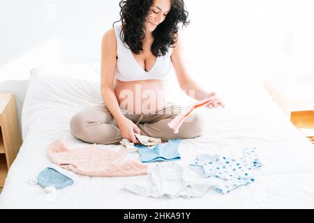 young pretty pregnant woman with curly black hair enjoys preparing the little clothes of her future baby sitting on the bed in her room. Stock Photo