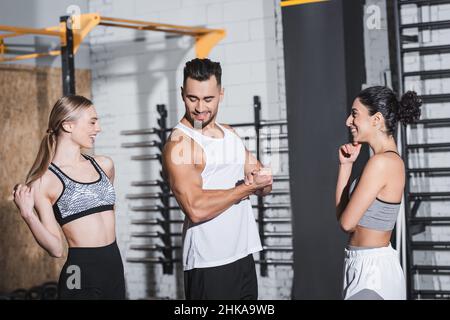 Smiling sportsman showing muscles near interracial sportswomen in gym Stock Photo