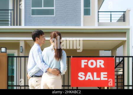Back portrait of Asian young couple standing and hugging together looking happy in front of their new house to start new life. Family, age, home, real Stock Photo