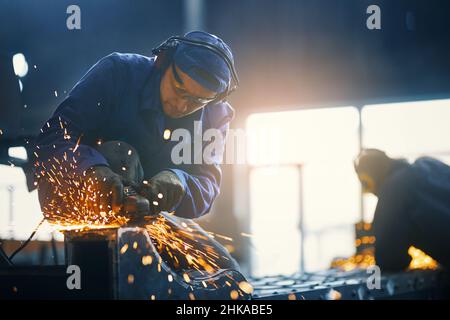 Caucasian man in protective clothes, glasses and gloves welding metal construction on factory. Professional locksmith making some details from steel at workshop.  Stock Photo