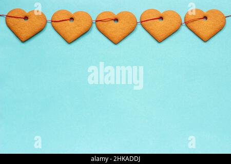 Garland of ginger cookies in shape of hearts on red ribbon, located on top of the frame edge on blue background. Top view Copy space for text, Valenti Stock Photo