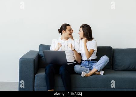 Overjoyed excited millennial couple getting good news, celebrating success, goal achieving, winning prize. Happy man and woman sitting on couch with l Stock Photo