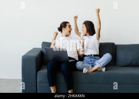 Overjoyed excited millennial couple getting good news, celebrating success, goal achieving, winning prize. Happy man and woman sitting on couch with l Stock Photo