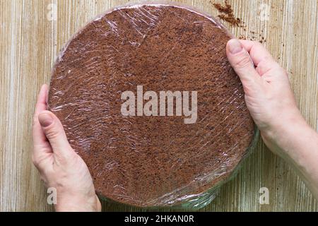 Chocolate sponge cake wrapped in cling film holding by woman hands on the wooden background. Top view Stock Photo