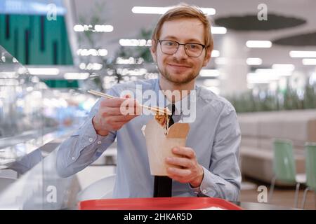 Portrait of smiling businessman with chopsticks eating chinese wok from box on food court. Lunch time. Stock Photo