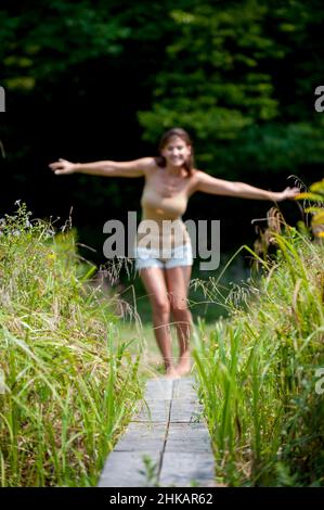 Young woman balancing on narrow wooden walkway, arms outstretched to help balance Stock Photo