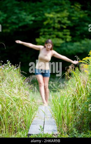 Young woman balancing on narrow wooden walkway, arms outstretched to help balance Stock Photo