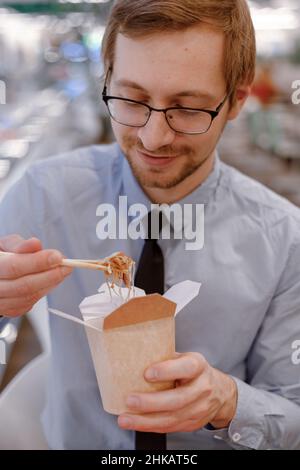 Smiling businessman in glasses eating chinese wok from box on food court. Lunch time. Stock Photo