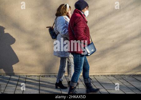 A woman leads an elderly lady wearing a Corona mask on a walkway by a wall of small mosaic stones. Behind them a shadow cast. Stock Photo