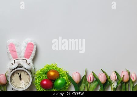 Easter concept. Alarm clock in the ears of a rabbit pearl eggs in a nest of flowers tulips on a gray background. copy space Stock Photo