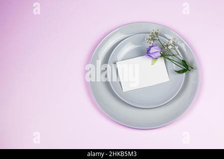 Holiday greeting mockup scene. Blank greeting cards, grey plates, and violet flowers eustoma branches on pink background with copy space. Flat lay, to Stock Photo