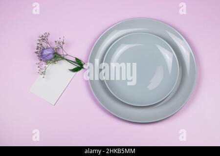 Holiday greeting mockup scene. Blank greeting cards, grey plates, and violet flowers eustoma branches on pink background with copy space. Flat lay, to Stock Photo