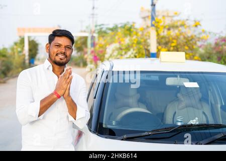Happy smiling cab driver greeting by doing namaste in front of car by looking at camera - concept of welcoming passenger, transportation service and Stock Photo