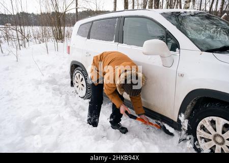 A man digs out a stalled car in the snow with a car shovel. Transport in winter got stuck in a snowdrift after a snowfall, sat on the bottom. First ai Stock Photo