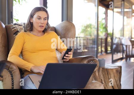 Caucasian pregnant woman using smartphone and laptop while working from home Stock Photo