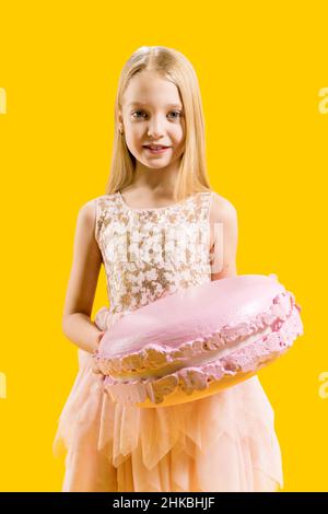 Christmas sweetness. Sweet gift. A cute girl is holding a huge pink macaroons on a yellow background. Round dessert. Stock Photo