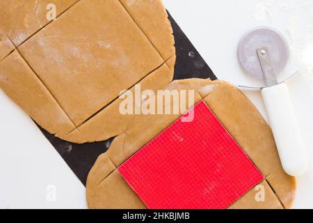 Cutting out walls of gingerbread house with knife for pizza on rolled raw gingerbread dough teflon baking sheet on white background Stock Photo