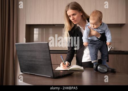 Young caucasian woman working at home in the office with a small baby. Mom holds her son in her arms, stands near the laptop and makes notes in a note Stock Photo