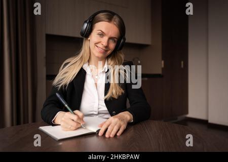 Smiling caucasian female student or freelancer in headphones writing in notepad in office, studying or working remotely. Lecture online wireless Stock Photo