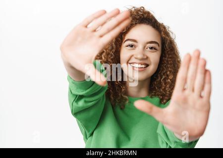 Portrait of happy beautiful girl, smiling, looking through finger frames, capturing moment, shooting photo, standing over white background Stock Photo