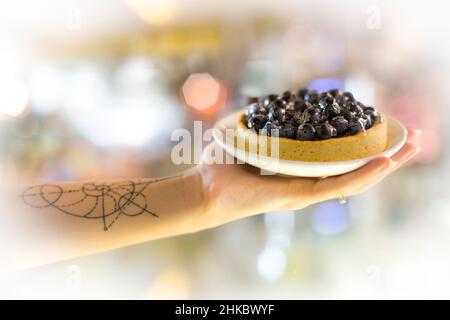 A tart with mascarpone and blueberries, in a female hand with a tattoo of a golden cross-section Stock Photo