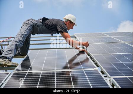 Worker mounting photovoltaic solar panel system outdoors. Man engineer placing solar module on metal rails, wearing construction helmets and work gloves. Renewable and ecological energy. Stock Photo