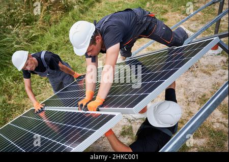 Male workers building photovoltaic solar panel system outdoors. Men engineers placing solar module on metal rails, wearing construction helmets and work gloves. Renewable and ecological energy. Stock Photo