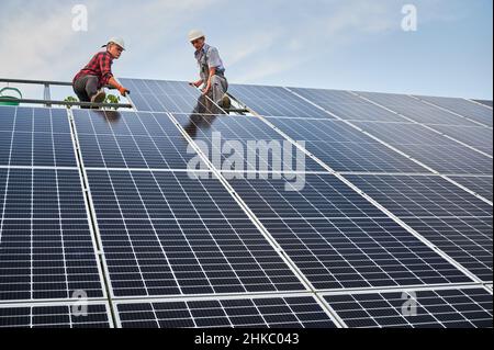 Low angle of two men solar technicians building photovoltaic solar panels under blue sky. Male workers in safety helmets placing solar module on metal rails. Concept of alternative energy sources. Stock Photo