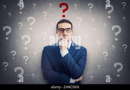 Skeptical businessman surrounded by interrogation marks has a main question as a red symbol over head. Thoughtful business person keeps hand under chi Stock Photo