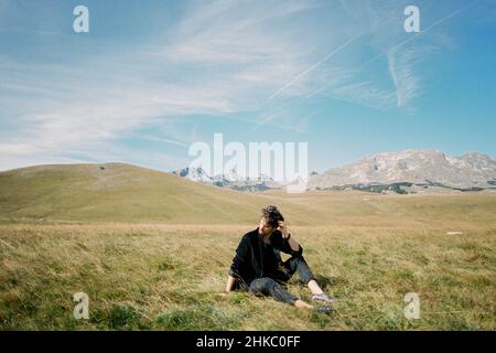 Man sits on a lawn against the backdrop of mountains, turning his head to the side Stock Photo