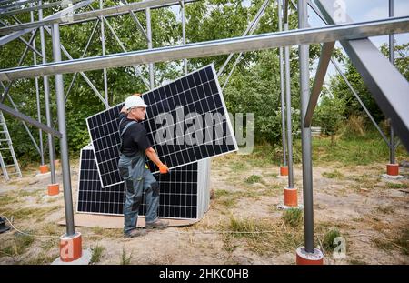 Full length of man solar installer carrying solar module while installing solar panel system in grassy area with trees. Male worker wearing safety helmet and work overalls while holding solar panel. Stock Photo