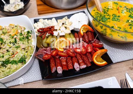 A black plate with sliced sausage, cucumbers and blue cheese lying on a table filled with food. Stock Photo