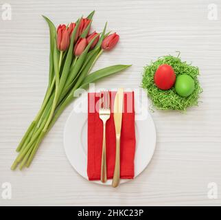 Easter table setting, Egg nest and red tulips decoration, gold cutlery and plate on white wooden background, top view. Spring Holiday. Stock Photo