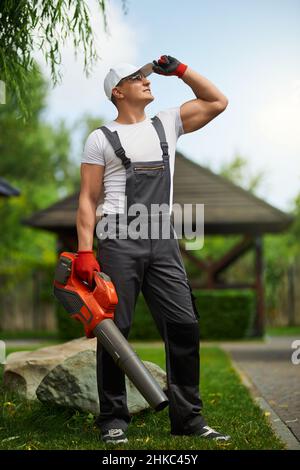 Full length portrait of handsome caucasian man in overalls, cap and gloves standing at garden with leaf blower in hands. Modern equipment for gardening. Stock Photo