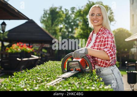 Beautiful female gardener in protective glasses and gloves trimming bushes with electric cutter. Charming woman with blond hair smiling and looking at camera at beautiful garden. Stock Photo