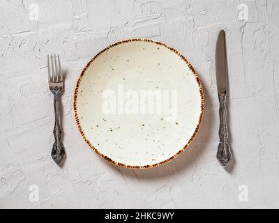 Set table. White speckled dinner plate between elegant table knife and fork over textured grey background. Empty porcelain plate with brown border. Stock Photo
