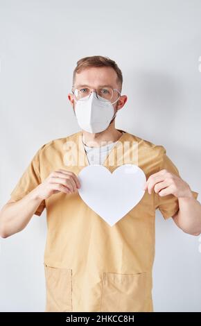 Portrait of caucasian male cardiologist doctor in medical uniform and medical mask with white paper heart shape. Adult doctor surgeon in hospital posing over the bright wall. High quality photo Stock Photo