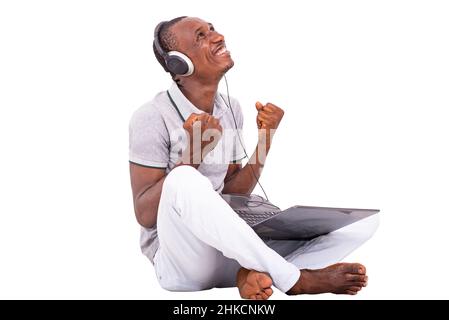 young man sitting on the floor working on laptop and listening to music smiling with raised fists. Stock Photo