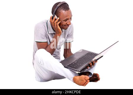 a handsome student in a t-shirt sitting on the floor isolated on a white background communicating while smiling using a laptop and headsets. Stock Photo