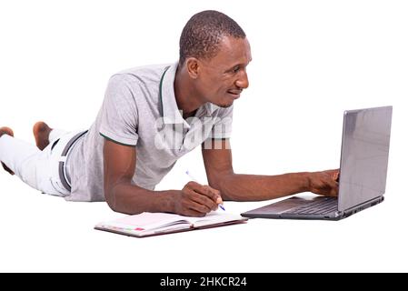 a handsome student in a t-shirt lying on the floor isolated on a white background working using a laptop taking notes while smiling. Stock Photo