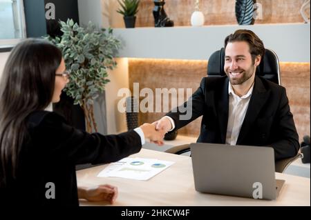 Successful young adult caucasian business colleagues, entrepreneurs, directors, sit at a desktop in modern office, made a good deal, agreed of cooperation, shake hands to each other, smiling Stock Photo