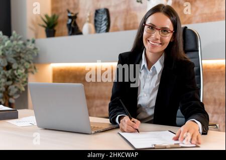 Portrait of a charming, intelligent Caucasian woman with glasses, office worker, a management company, trader investor, sit at work desk in modern office, in stylish business clothes, smiling Stock Photo
