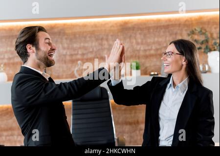 Positive successful young adult Caucasian business colleagues, managers, company employees, bank coworkers, finished the working day, closed an important deal, get profit,are giving five to each other Stock Photo